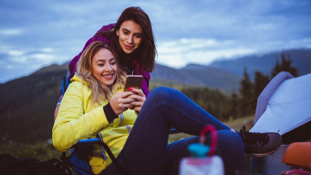 NorthSiginal Two women outdoors at a campsite, one sitting in a chair using her smartphone with North Signal Wifi Rental while the other leans over her shoulder, both smiling with mountains and trees in the background. Starlink Rental