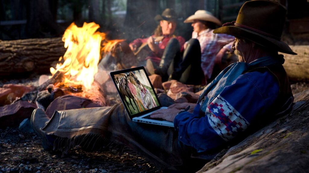 NorthSiginal A person in cowboy attire uses a laptop near a campfire, powered by North Signal Wifi Rental, while two others sit in the background; the forest setting is visible around them. Starlink Rental