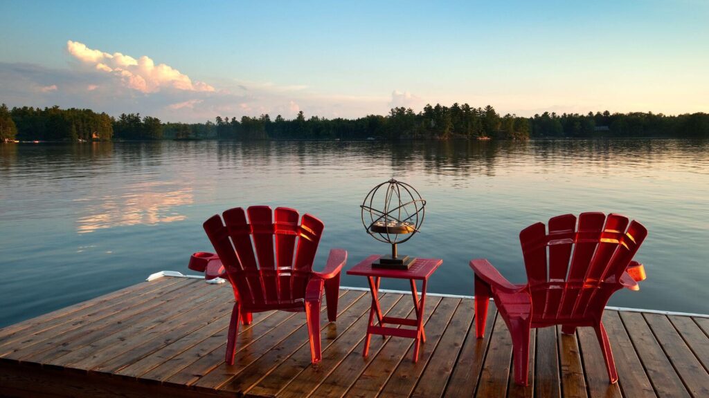 NorthSiginal Two red Adirondack chairs and a small table sit on a wooden dock overlooking a calm lake at sunset, with North Signal Wifi Rental ensuring you stay connected as you relax amidst trees and clouds in the distance. Starlink Rental